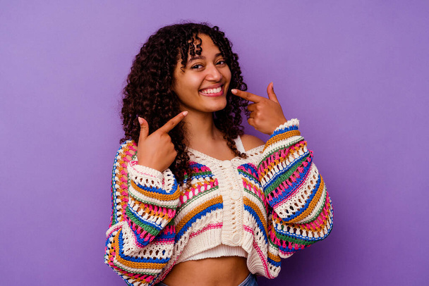 Young mixed race woman isolated on purple background smiles, pointing fingers at mouth. - Fotoğraf, Görsel