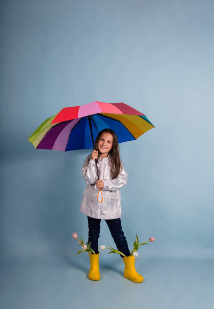 une jolie petite fille brune en bottes en caoutchouc avec des fleurs se tient sous un parapluie multicolore sur un fond bleu avec une place pour le texte - Photo, image