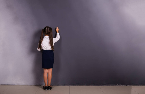 Μια μαθήτρια με στολή στέκεται με την πλάτη της σε ένα φόντο με μια πλάκα. - Φωτογραφία, εικόνα