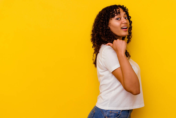 Jonge gemengde ras vrouw geïsoleerd op gele achtergrond punten met duim vinger weg, lachen en zorgeloos. - Foto, afbeelding