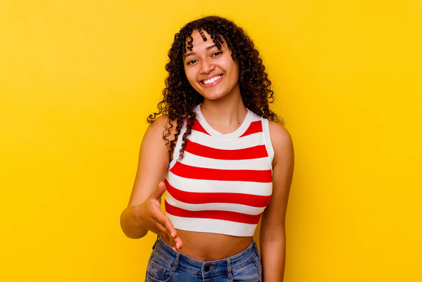 Young mixed race woman isolated on yellow background stretching hand at camera in greeting gesture. - Photo, Image