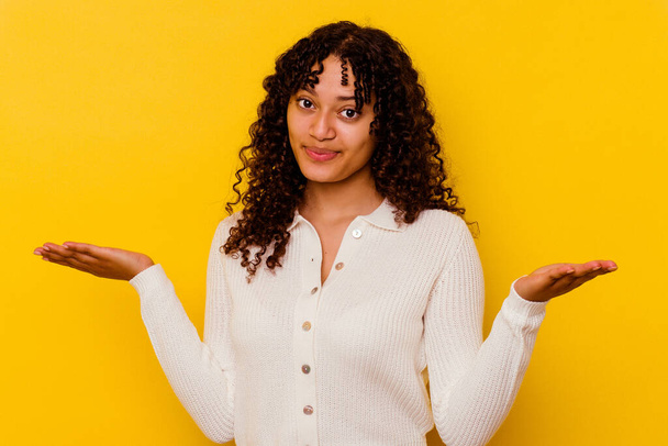 Jonge gemengde ras vrouw geïsoleerd op gele achtergrond twijfelen en schouders ophalen in vragen gebaar. - Foto, afbeelding