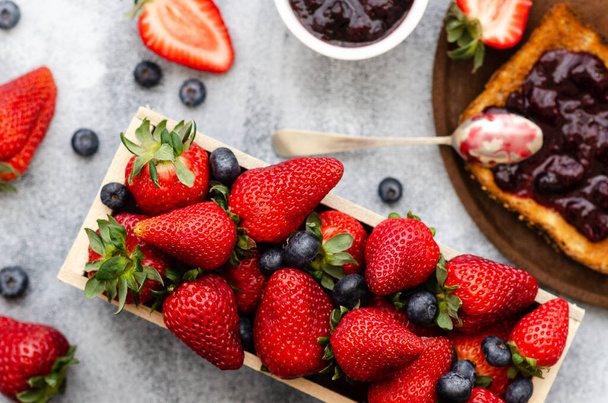 Erdbeeren und Blaubeeren in einer rechteckigen Holzkiste, eine Scheibe Toast mit Erdbeer- und Blaubeermarmelade auf einem Holzteller und einem Löffel und Erdbeer- und Blaubeermarmelade in einer weißen Schüssel.  - Foto, Bild