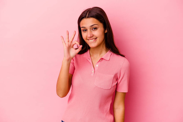 Junge Indianerin, isoliert auf rosa Hintergrund, zwinkert und hält eine Okay-Geste mit der Hand. - Foto, Bild
