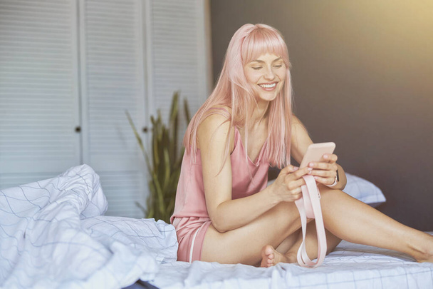 Ευτυχισμένη κυρία με μακριά ροζ μαλλιά κρατά smartphone κάθεται σε άνετο κρεβάτι - Φωτογραφία, εικόνα
