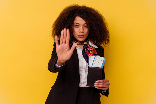 Młoda afrykańska stewardesa trzymająca bilety lotnicze na żółtym tle, stojąca wyciągniętą ręką i pokazująca znak stopu.. - Zdjęcie, obraz