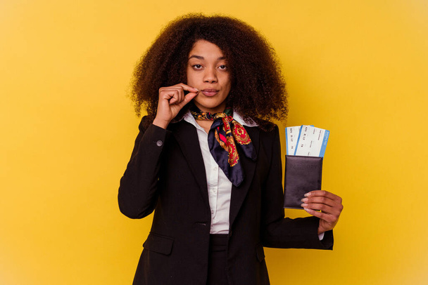Młoda afrykańska stewardesa trzymająca bilety na samolot odizolowane na żółtym tle z palcami na ustach trzymającymi sekret. - Zdjęcie, obraz
