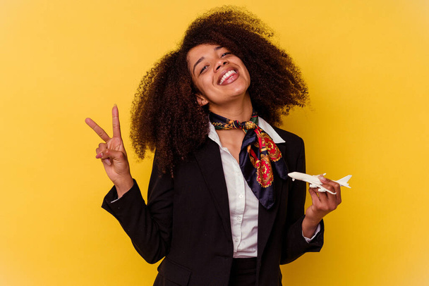 Giovane hostess aerea afroamericana che tiene un piccolo aereo isolato su sfondo giallo gioioso e spensierato mostrando un simbolo di pace con le dita. - Foto, immagini