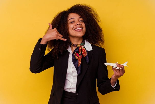Νεαρή Αφροαμερικάνα αεροσυνοδός που κρατά ένα μικρό αεροπλάνο απομονωμένο σε κίτρινο φόντο που δείχνει μια χειρονομία κινητής τηλεφωνίας με δάχτυλα. - Φωτογραφία, εικόνα