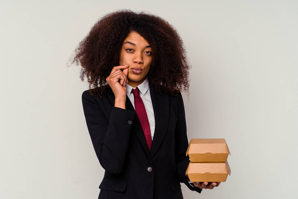 Jovem mulher de negócios afro-americana segurando um hambúrguer isolado no fundo branco com os dedos nos lábios mantendo um segredo. - Foto, Imagem