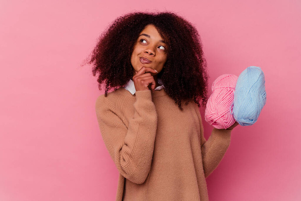 Junge afrikanisch-amerikanische Frau hält einen Nähfaden isoliert auf rosa Hintergrund und blickt mit zweifelndem und skeptischem Gesichtsausdruck zur Seite. - Foto, Bild