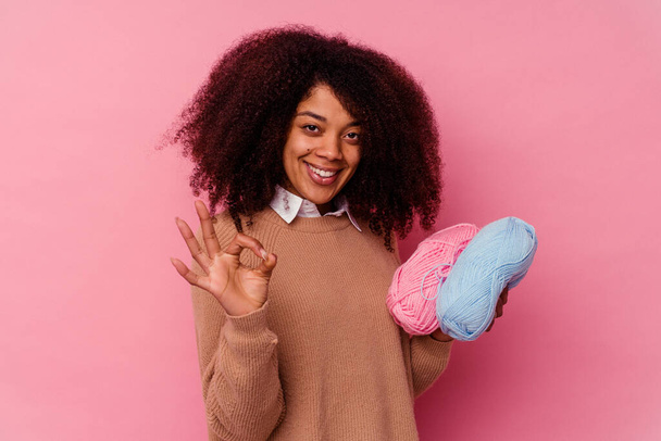 Joven mujer afroamericana sosteniendo un hilo de coser aislado sobre fondo rosa alegre y confiado mostrando gesto ok. - Foto, imagen