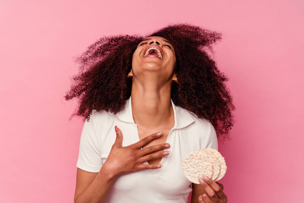Νεαρή Αφροαμερικανή που τρώει κέικ ρυζιού απομονωμένη σε ροζ φόντο γελάει δυνατά κρατώντας το χέρι στο στήθος. - Φωτογραφία, εικόνα