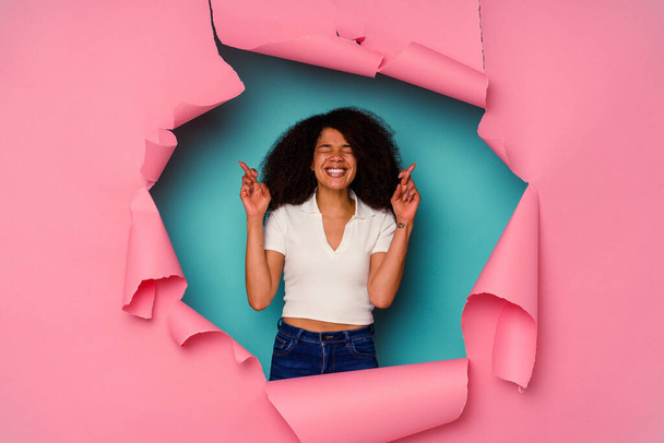 Junge Afroamerikanerin in zerrissenem Papier isoliert auf blauem Hintergrund drückt Glück die Daumen - Foto, Bild