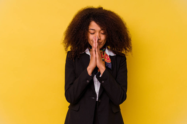Giovane hostess aerea afroamericana isolata su sfondo giallo tenendosi per mano in preghiera vicino alla bocca, si sente fiducioso. - Foto, immagini