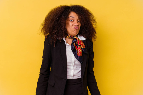 Giovane hostess aerea afroamericana isolata su sfondo giallo scrolla spalle e occhi aperti confusi. - Foto, immagini