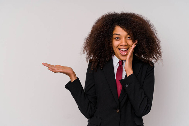 Young African American business woman wearing a suit isolated on white background holds copy space on a palm, keep hand over cheek. Amazed and delighted. - Photo, Image