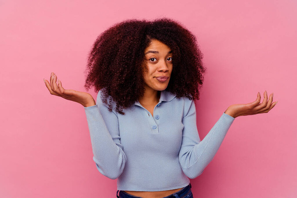 Jeune femme afro-américaine isolée sur fond rose épaules confuses et douteuses haussant pour tenir un espace de copie. - Photo, image
