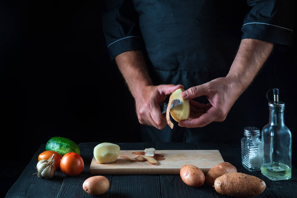 Profi-Koch schält Kartoffeln für Pommes. Für Kartoffelgerichte. Auf schwarzem Hintergrund Konzept für Speisekarte, Kochen, gesunde Ernährung. Freie Werbefläche - Foto, Bild