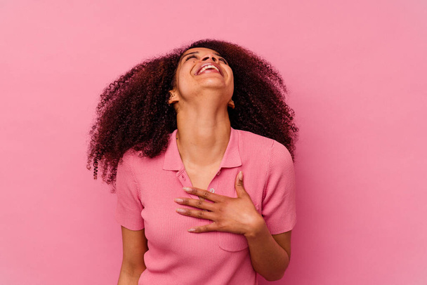 Mujer afroamericana joven aislada sobre fondo rosa se ríe en voz alta manteniendo la mano en el pecho. - Foto, imagen