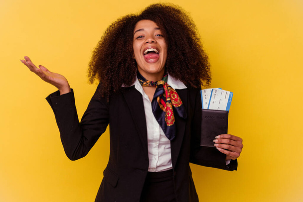 Genç Afrikalı Amerikalı hostes sarı arka planda izole edilmiş uçak biletlerini tutuyor. Hoş bir sürpriz, heyecan verici ve el kaldırıyor.. - Fotoğraf, Görsel