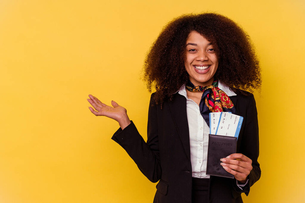 Young african american air hostess holding a plane tickets isolated on yellow background  showing a copy space on a palm and holding another hand on waist. - Foto, afbeelding