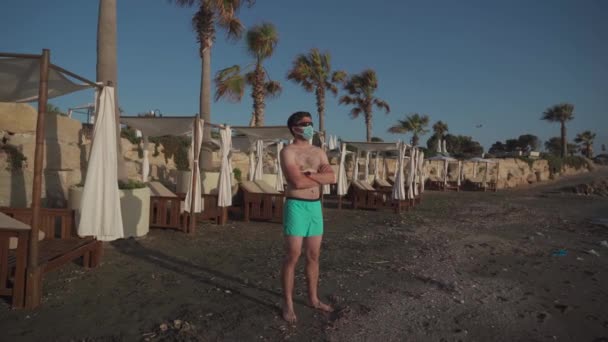 ヤシの木やシラキュースの地中海沿岸のサンラウンジャーを背景に、汚れたビーチだけで保護マスクの男を休ませます。男性持っています夏休み中にcovid lockdown - 映像、動画