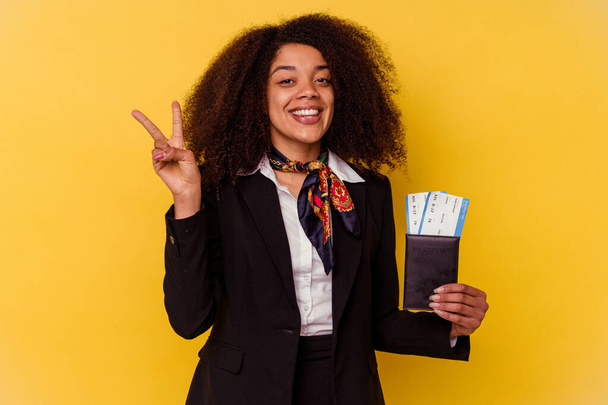 Jeune hôtesse de l'air afro-américaine tenant un billet d'avion isolé sur fond jaune montrant le numéro deux avec les doigts. - Photo, image