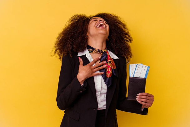 Jeune hôtesse de l'air afro-américaine tenant un billet d'avion isolé sur fond jaune rit bruyamment en gardant la main sur la poitrine. - Photo, image