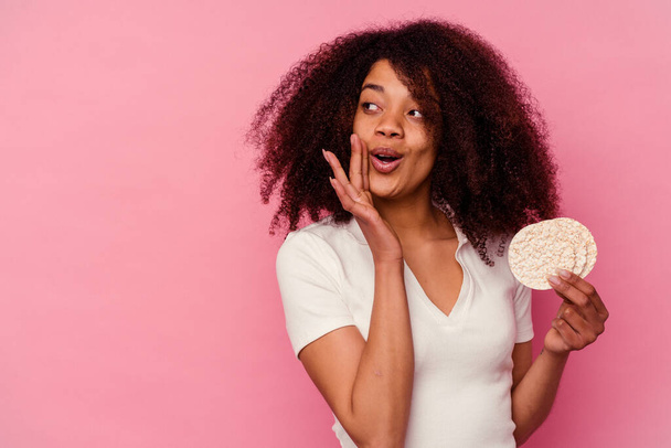 Junge afrikanisch-amerikanische Frau isst einen Reiskuchen isoliert auf rosa Hintergrund sagt eine geheime heiße Bremse Nachrichten und Blick zur Seite - Foto, Bild