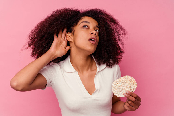 Joven mujer afroamericana comiendo pasteles de arroz aislados sobre fondo rosa tratando de escuchar un chisme. - Foto, imagen