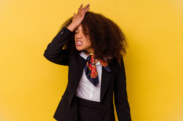 Νεαρή Αφρο-Αμερικανίδα αεροσυνοδός απομονωμένη σε κίτρινο φόντο ξεχνώντας κάτι, χτυπώντας το μέτωπο με παλάμη και κλείνοντας τα μάτια. - Φωτογραφία, εικόνα