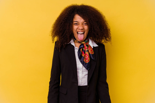 Joven afro-americana azafata aislada sobre fondo amarillo divertida y amigable sobresaliendo lengua. - Foto, imagen