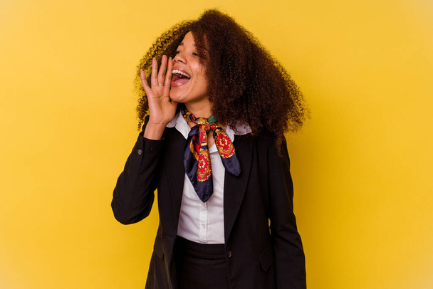 Νεαρή Αφρο-Αμερικανίδα αεροσυνοδός απομονωμένη σε κίτρινο φόντο φωνάζοντας και κρατώντας παλάμη κοντά στο ανοιχτό στόμα. - Φωτογραφία, εικόνα