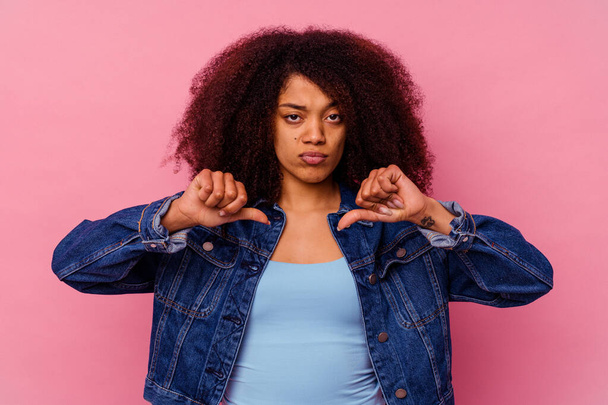 Jonge Afro-Amerikaanse vrouw geïsoleerd op roze achtergrond toont een afkeer gebaar, duimen naar beneden. Concept van onenigheid. - Foto, afbeelding