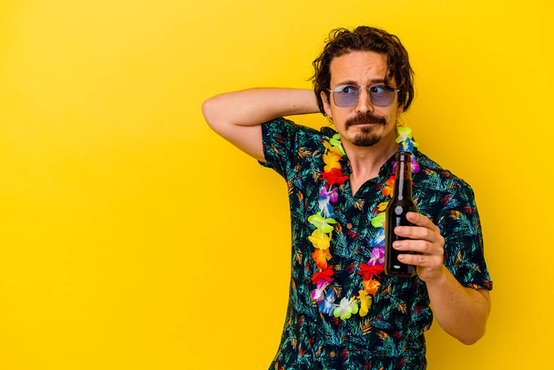 Jonge blanke man draagt een hawaiiaanse ketting met een bier geïsoleerd op een gele achtergrond die de achterkant van het hoofd raakt, nadenkt en een keuze maakt. - Foto, afbeelding