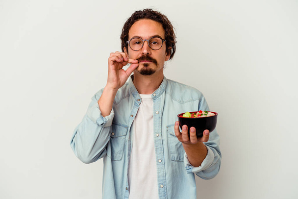 Junger kaukasischer Mann isst einen Ramen isoliert auf weißem Hintergrund mit Fingern auf den Lippen, der ein Geheimnis bewahrt. - Foto, Bild