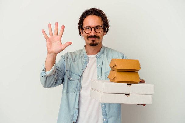 Young caucasian man holding hamburger an pizzas isolated on white background smiling cheerful showing number five with fingers. - Valokuva, kuva