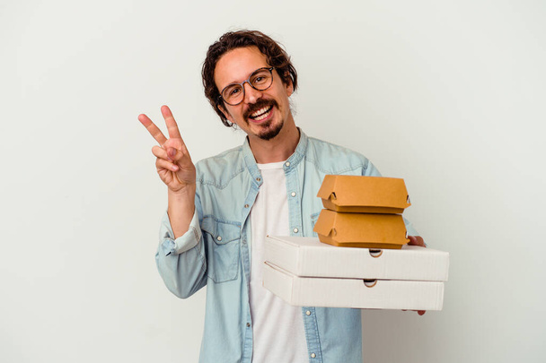 若い白人男性がハンバーガーを持っている白い背景に隔離されたピザ喜びと屈託のない指で平和のシンボルを示す. - 写真・画像