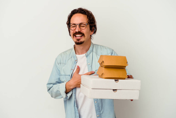 Jovem caucasiano segurando hambúrguer uma pizzas isolado no fundo branco ri alto mantendo a mão no peito. - Foto, Imagem
