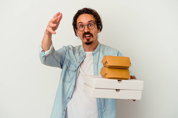 Jeune homme caucasien tenant hamburger une pizzas isolées sur fond blanc recevant une agréable surprise, excité et levant les mains. - Photo, image