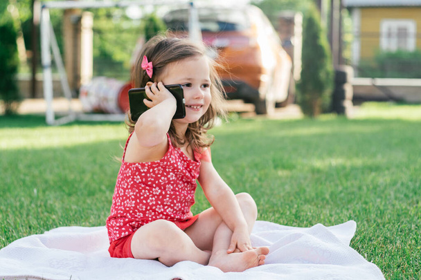 Mała 3-4 dziewczyna w czerwonych ubraniach siedzi na kocu na zielonej trawie i mówi przez telefon komórkowy. Dzieci, za pomocą gadżetów - Zdjęcie, obraz