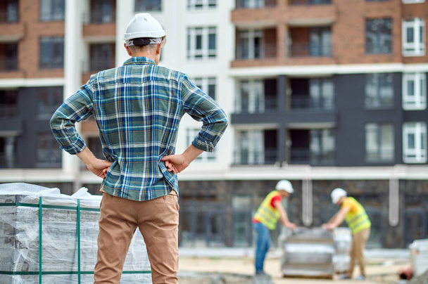 Civil engineer in protective headwear looking at two male builders - Foto, afbeelding