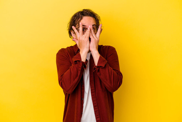 Young caucasian man isolated on yellow background blink at the camera through fingers, embarrassed covering face. - Foto, imagen