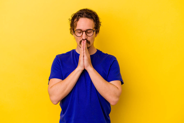 Young caucasian man isolated on yellow background praying, showing devotion, religious person looking for divine inspiration. - Fotografie, Obrázek