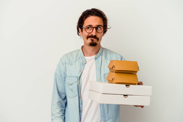 Junge kaukasische Mann hält Hamburger und Pizzen isoliert auf weißem Hintergrund verwirrt, fühlt sich zweifelhaft und unsicher. - Foto, Bild