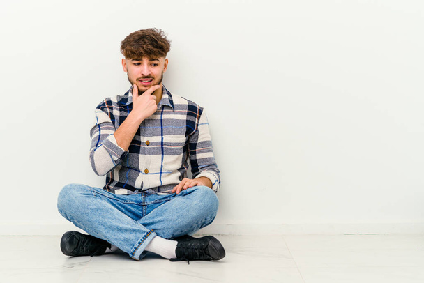 Молодий марокканець сидить на підлозі, ізольований на білому фоні, торкаючись потилиці, думаючи і роблячи вибір.. - Фото, зображення