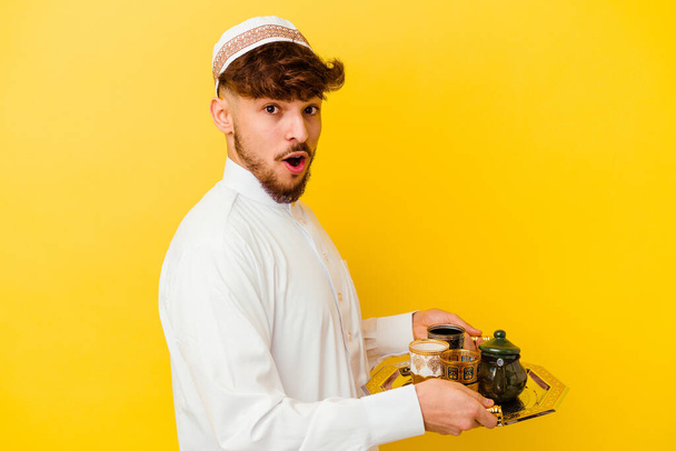 Young Moroccan man wearing the typical arabic costume drinking tea isolated on yellow background - Photo, Image