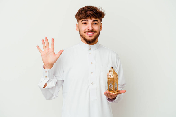 Junger marokkanischer Mann in typischer arabischer Kleidung hält eine Ramadanlampe auf weißem Hintergrund und lächelt fröhlich und zeigt Nummer fünf mit den Fingern. - Foto, Bild