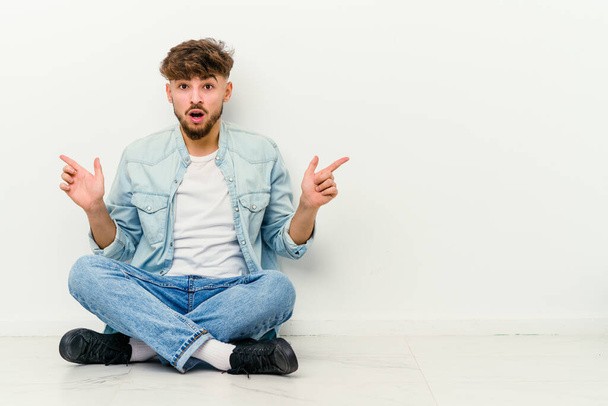 Young Moroccan man sitting on the floor isolated on white background pointing to different copy spaces, choosing one of them, showing with finger. - Photo, Image